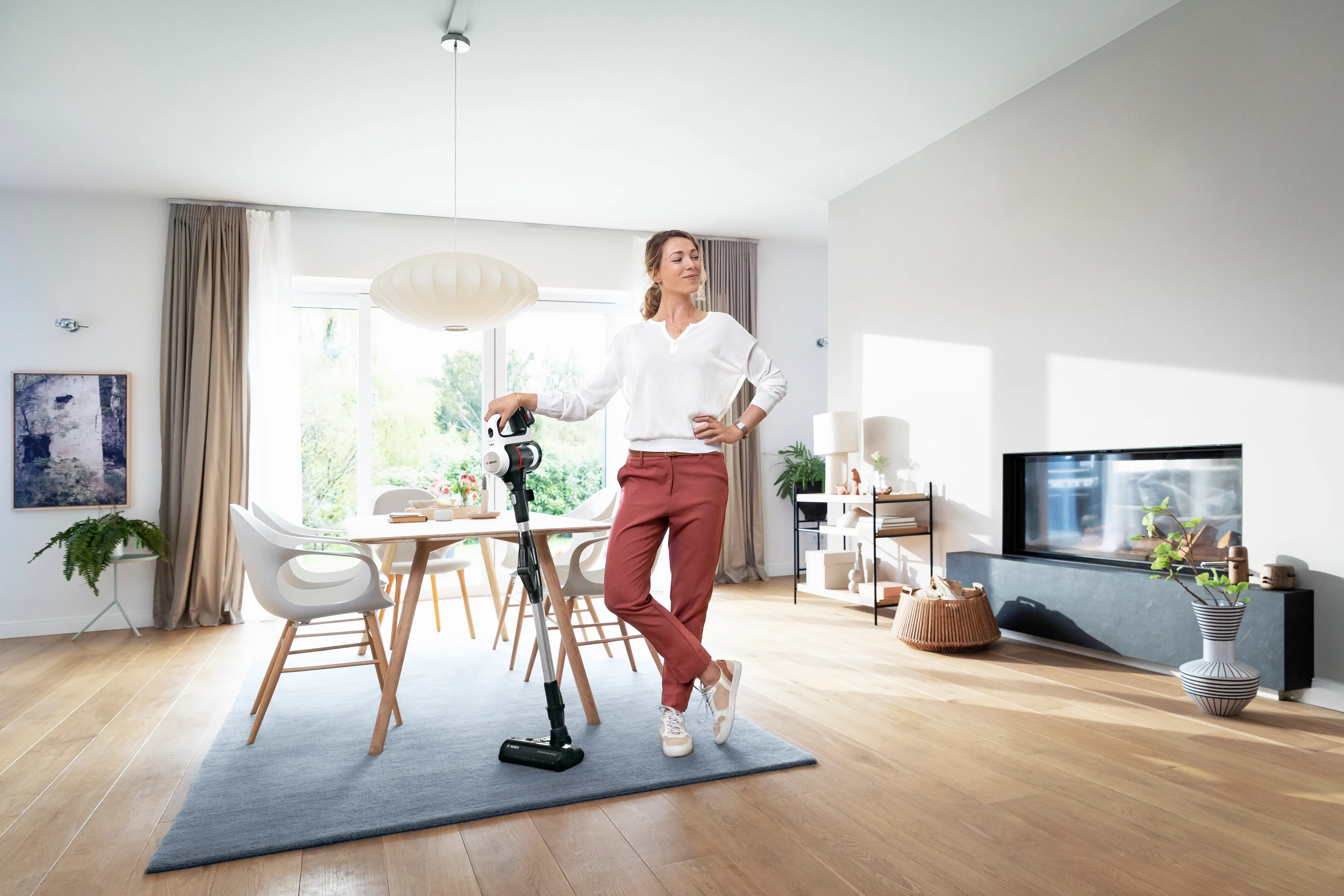 A woman stands confidently beside a Bosch cordless vacuum cleaner in a bright, modern living and dining room, suggesting effortless cleaning.