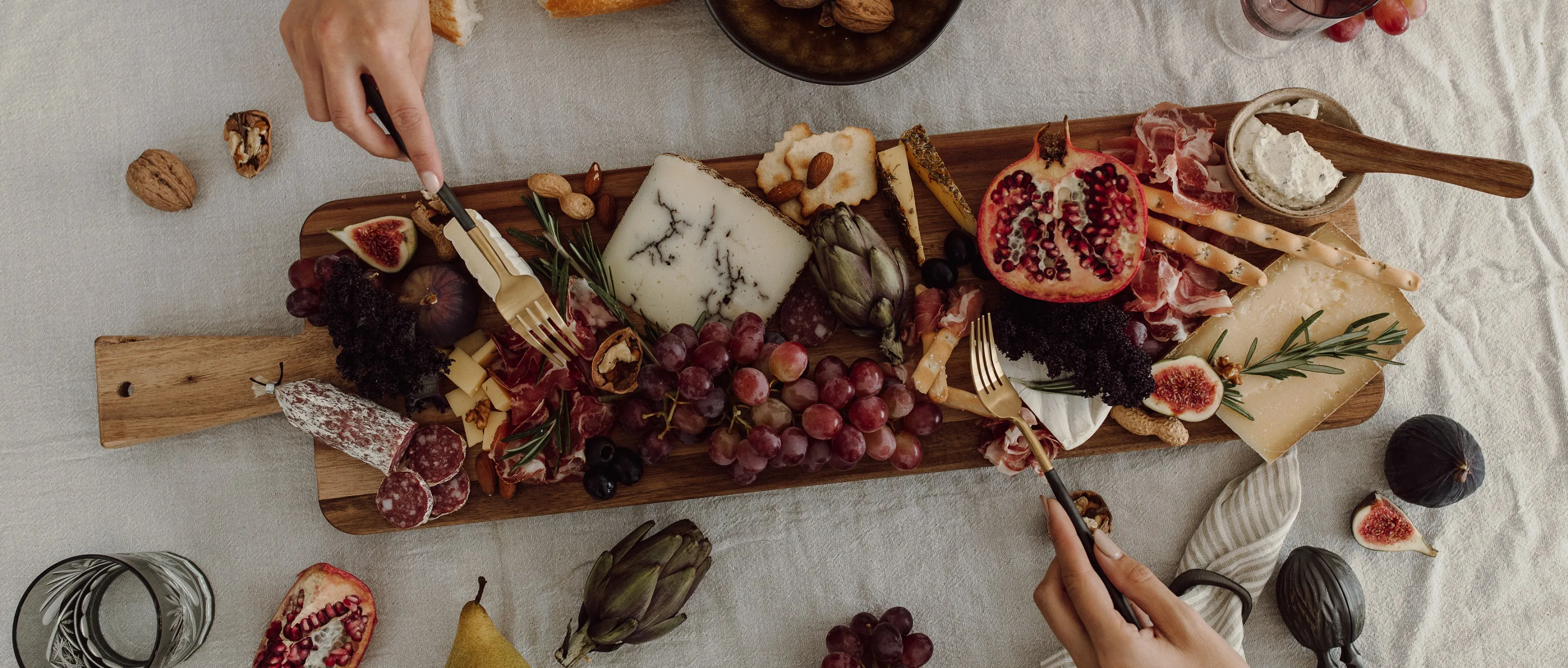 Ein Holzbrett mit Käse, Wurst, Früchten und Nüssen, eine Hand wählt mit einer Gabel ein Stück aus.