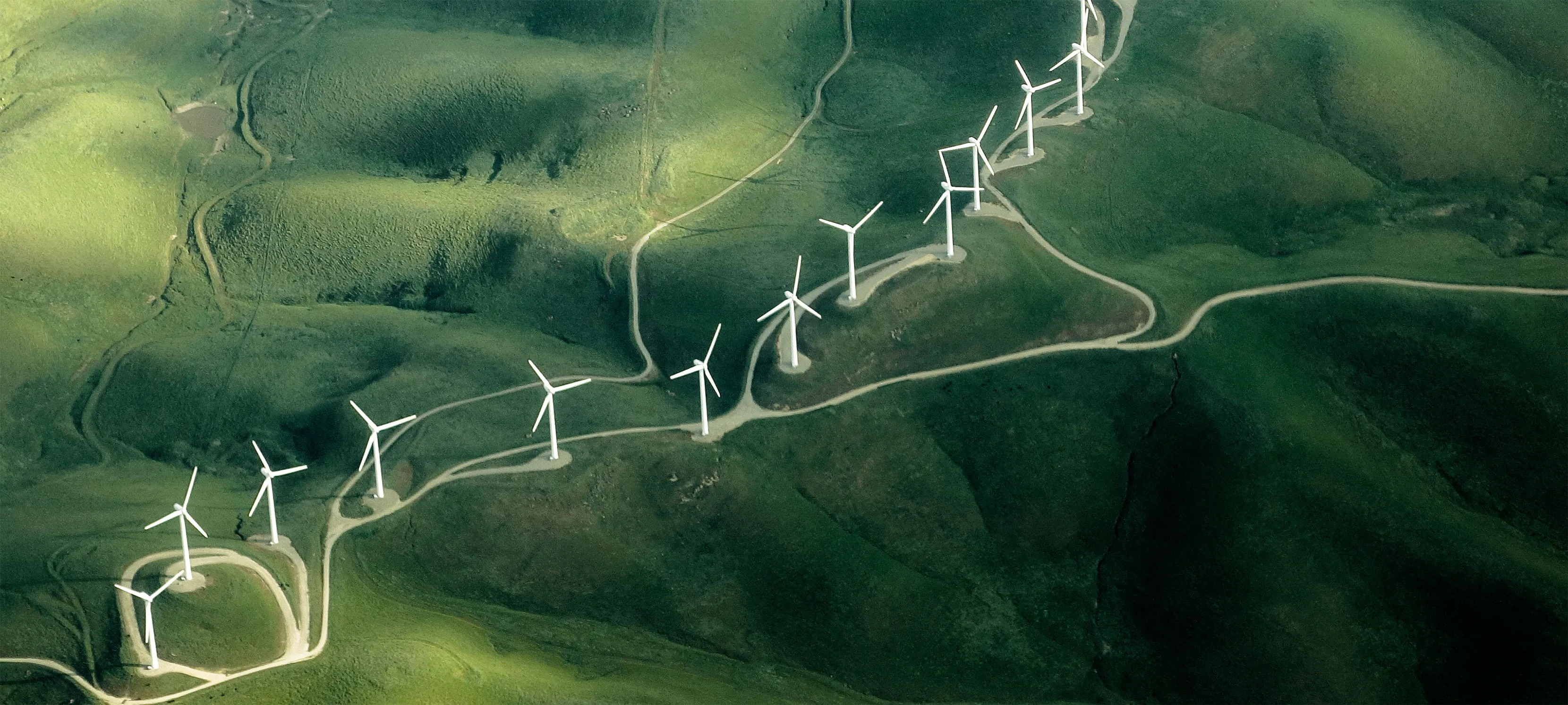 Aerial view of wind turbines arranged along a winding road amidst green hills.