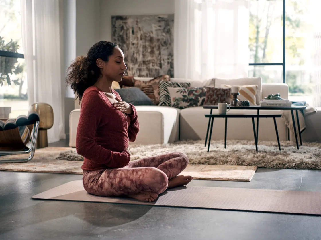 Woman in red sweater meditating on yoga mat in living room with couch and table.