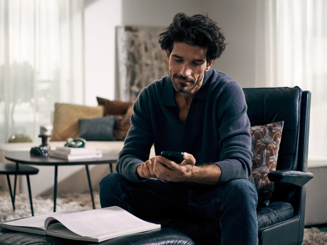 Man with curly hair sitting in armchair, reading book, surrounded by home decor.