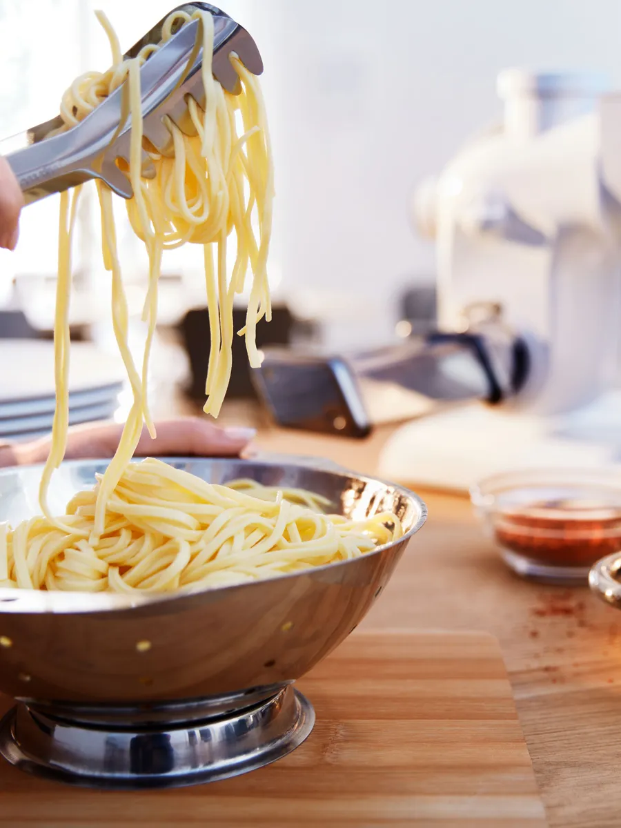 A hand uses tongs to lift freshly cooked spaghetti from a stainless steel bowl, with a pasta machine and ingredients visible in the background.