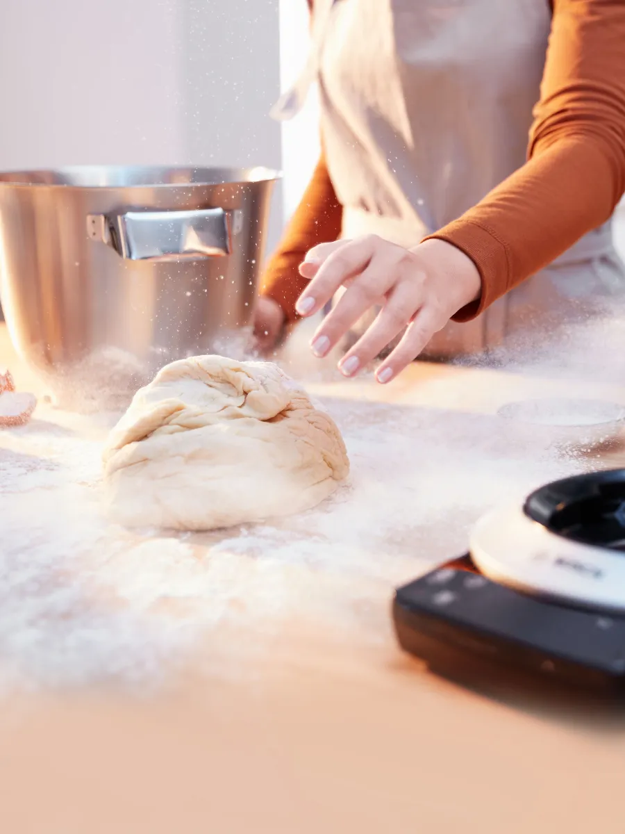 A person in an apron sprinkles flour over a ball of dough on a wooden surface, with a metal mixing bowl and a waffle maker nearby.