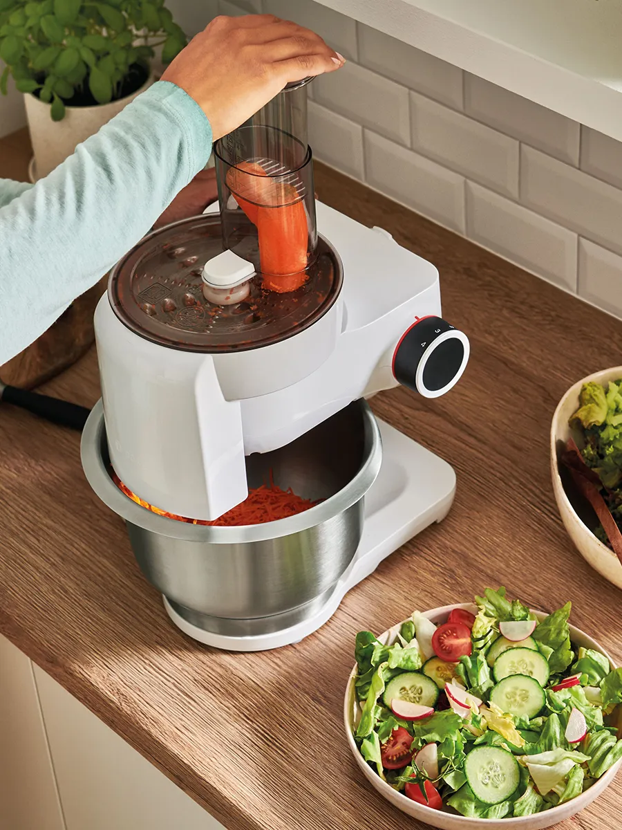 A person using a food processor to shred carrots, with a fresh salad nearby on a wooden countertop.