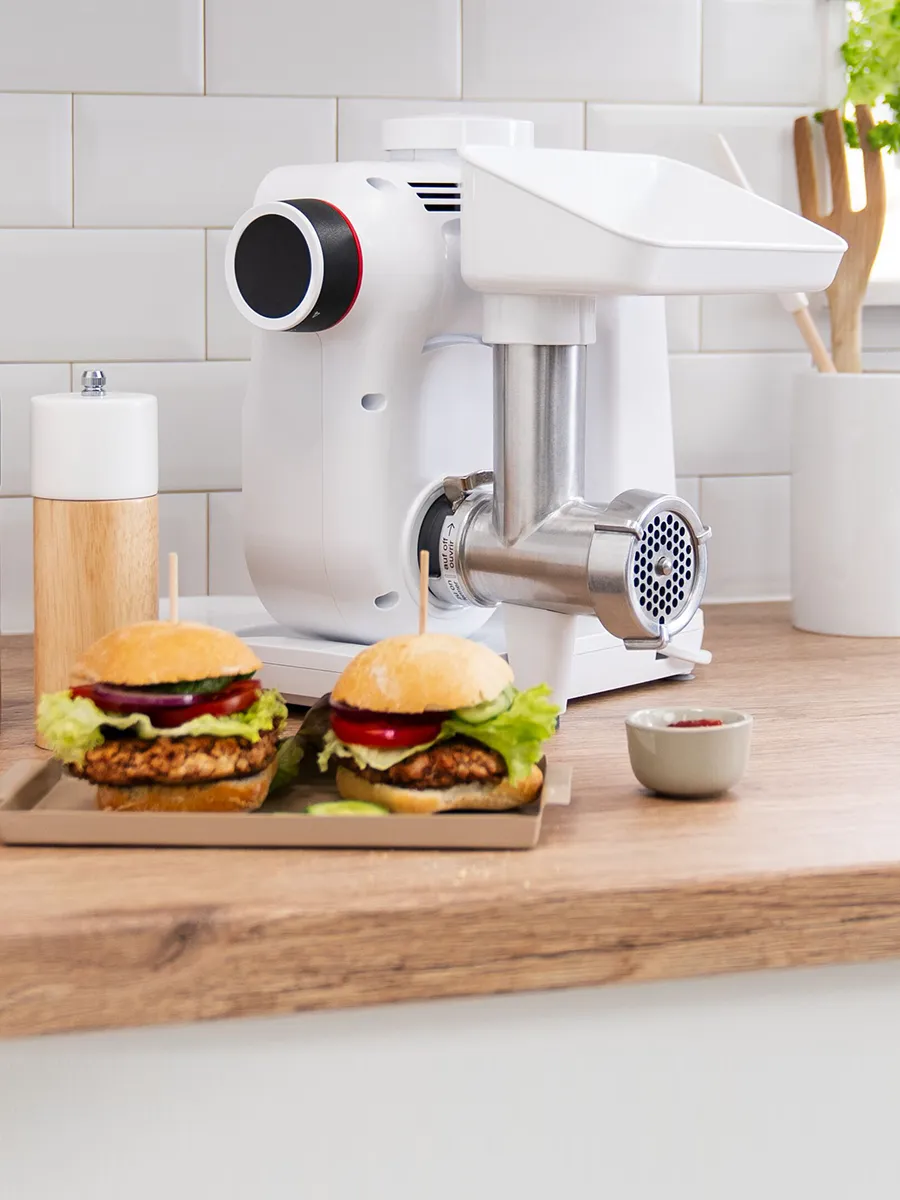 A modern meat grinder on a kitchen counter, with two crispy chicken sliders topped with lettuce and ketchup in the foreground.