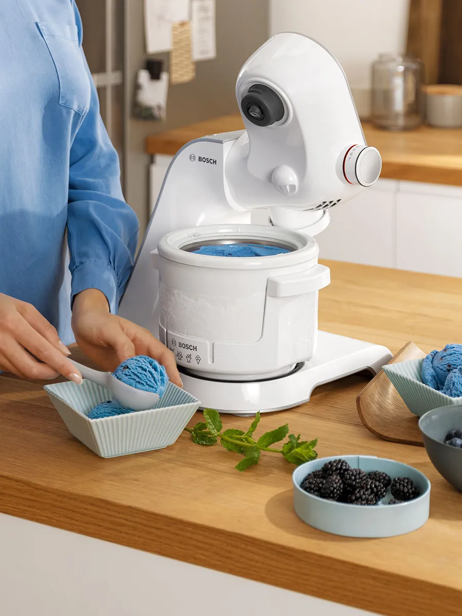 A person scoops blue ice cream from a Bosch ice cream maker into bowls on a wooden kitchen counter, surrounded by mint and blackberries.