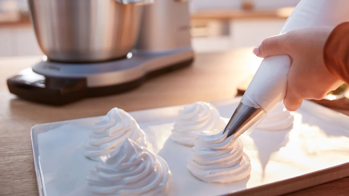 Hand holding a piping bag, piping swirled white meringue or whipped cream onto a baking tray in a sunlit kitchen with a stand mixer in the background.