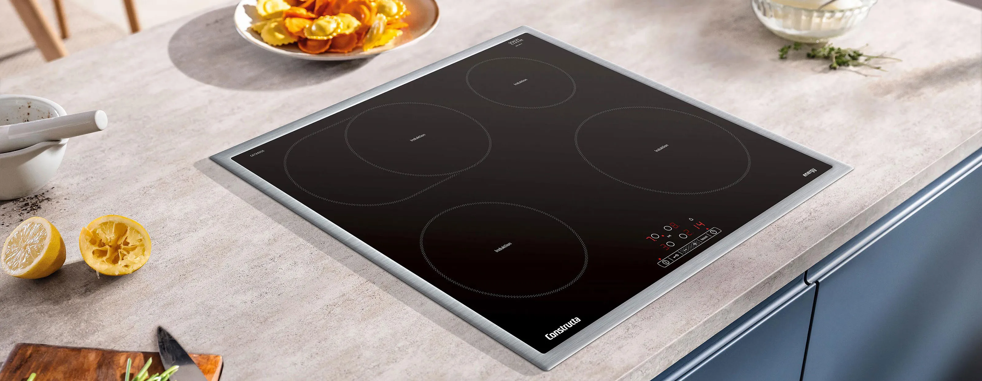 Ein schlanker schwarzer Induktionskochfeld mit vier Platten, auf einer hellen Steinarbeitsplatte, umgeben von Küchenutensilien und frischen Zutaten.