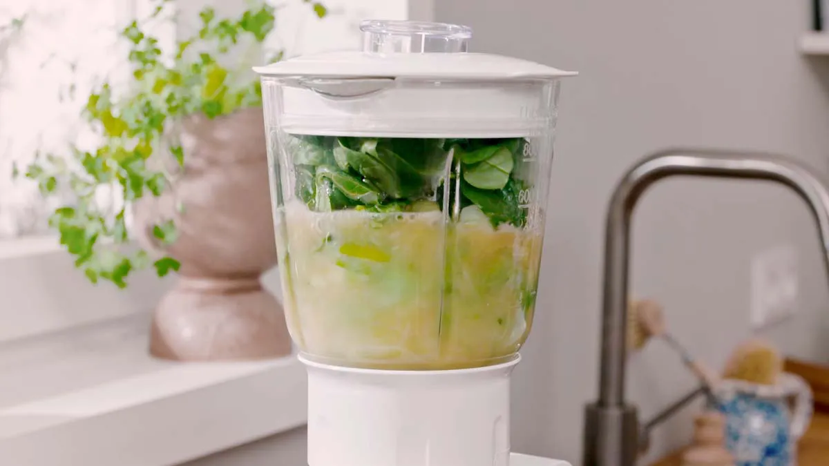 Blender with green leafy vegetables and liquid, kitchen counter, window, and other household items.