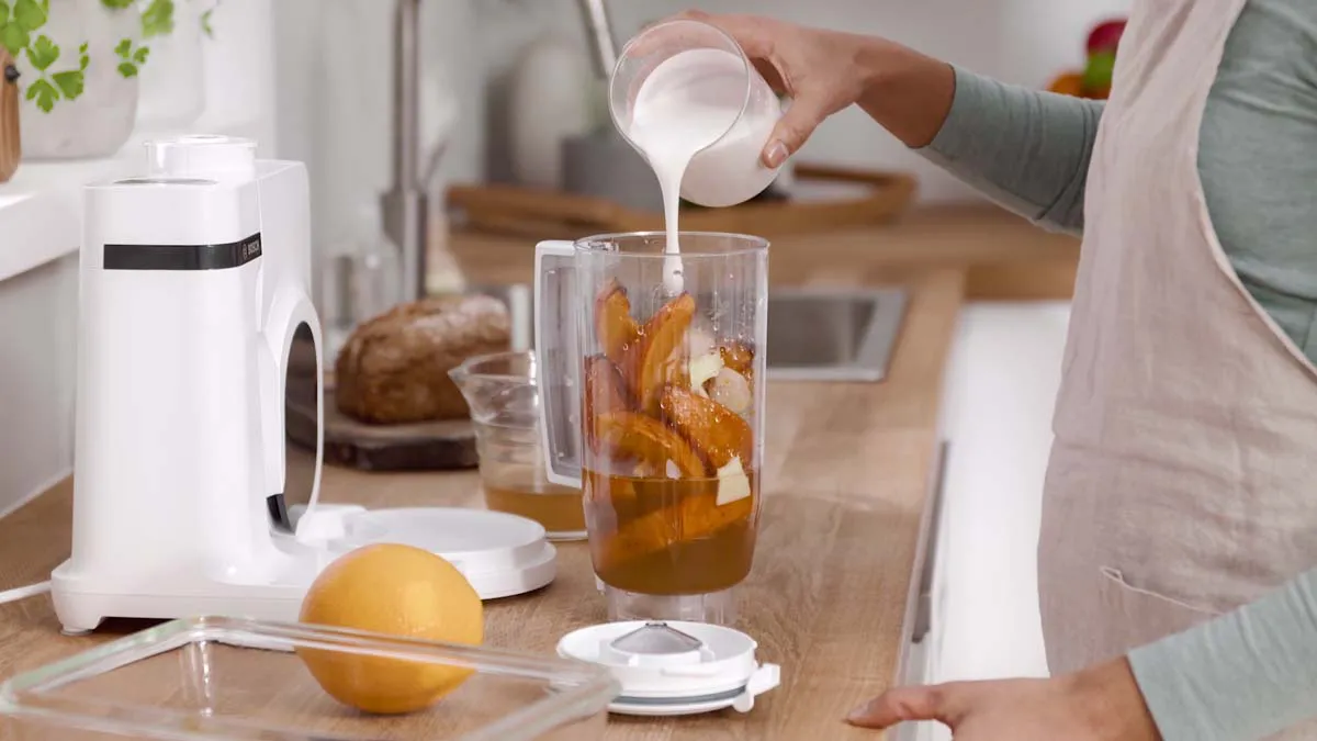 Person pouring milk into a glass containing orange slices and other ingredients on a kitchen counter.