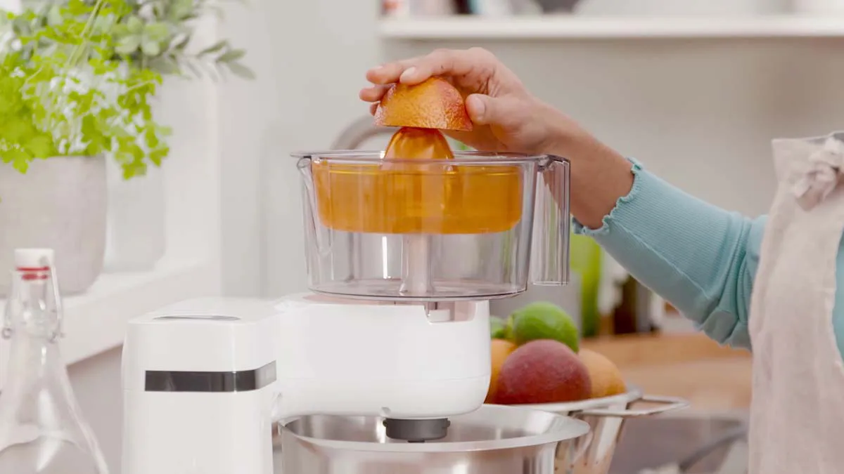 Hands squeezing orange into juicer, kitchen counter with fresh produce, modern appliances.