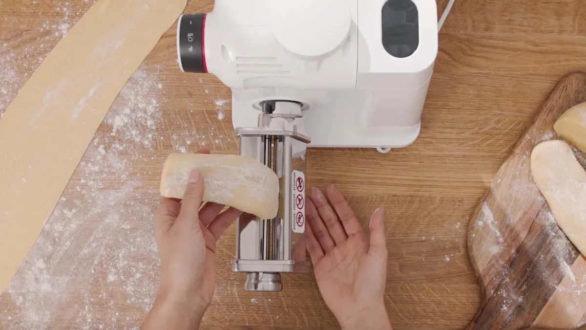 Hands using a pasta maker machine on a wooden surface with flour.