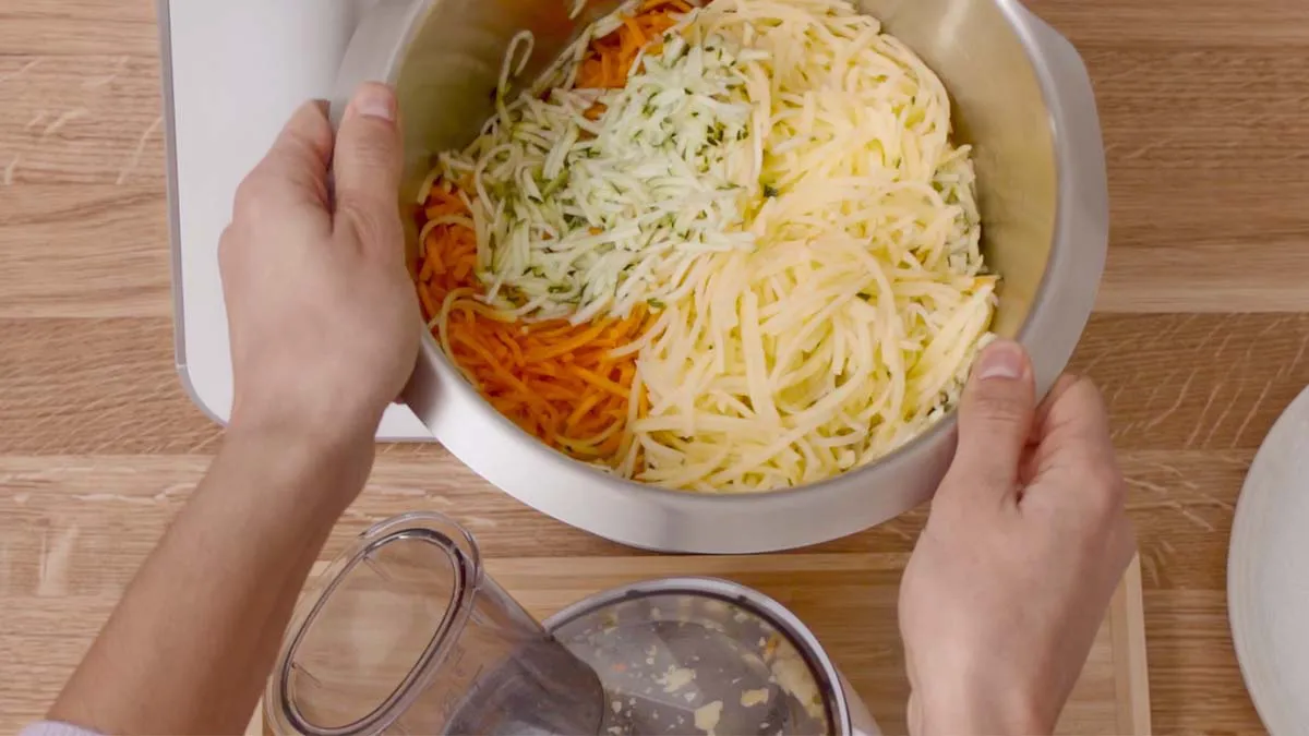 Bowl of shredded carrots and cheese on wooden table, hands adding ingredients.
