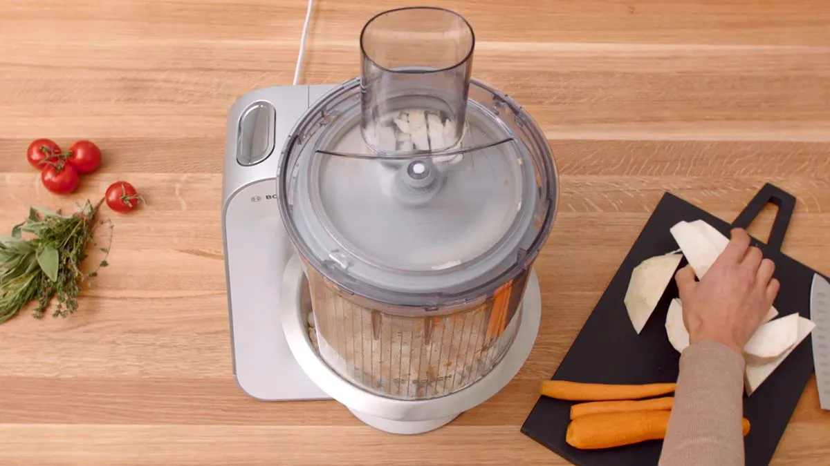 Food processor with vegetables, kitchen utensils, and wooden surface.