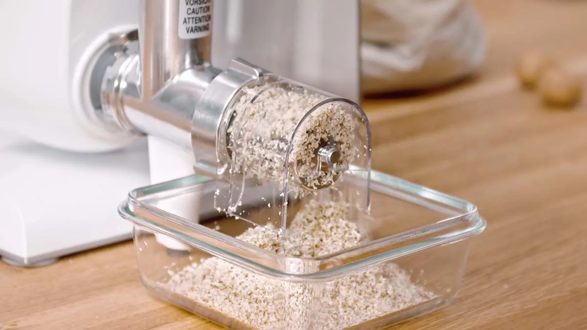 Electric grain mill grinding oats into a glass container on a wooden surface.