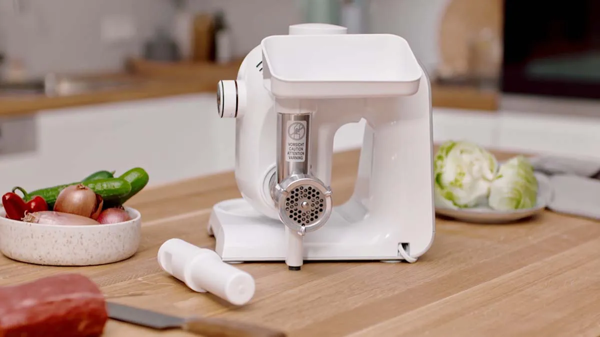 White electric meat grinder on wooden table with fresh vegetables.