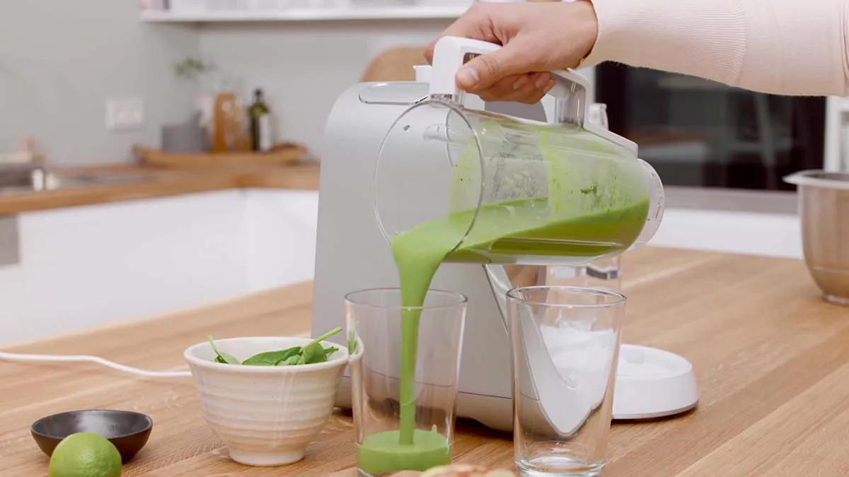 A person pouring green liquid from a blender into a glass on a kitchen counter.