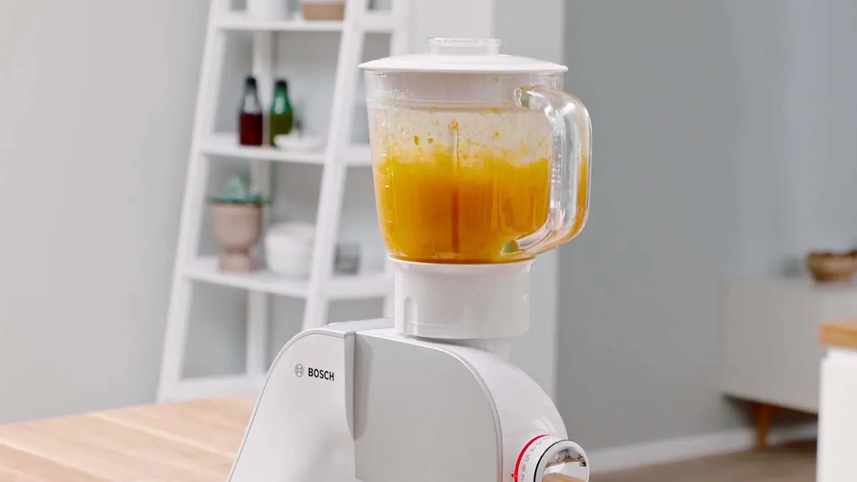 Blender with orange liquid on kitchen counter, shelves in background.