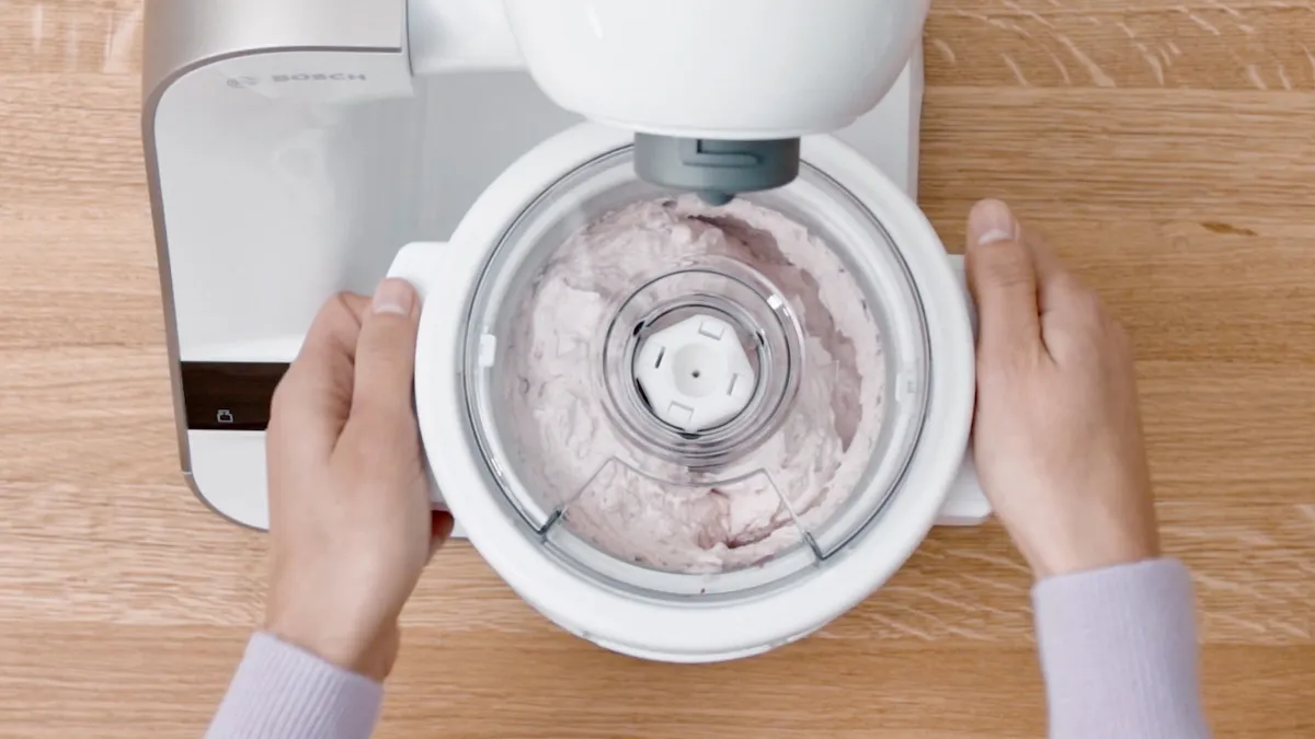 Hands operating a food processor, mixing ingredients on a wooden table.