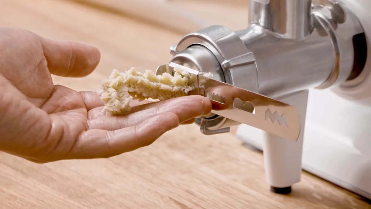 Hands operating a meat grinder to process food ingredients on a wooden surface.