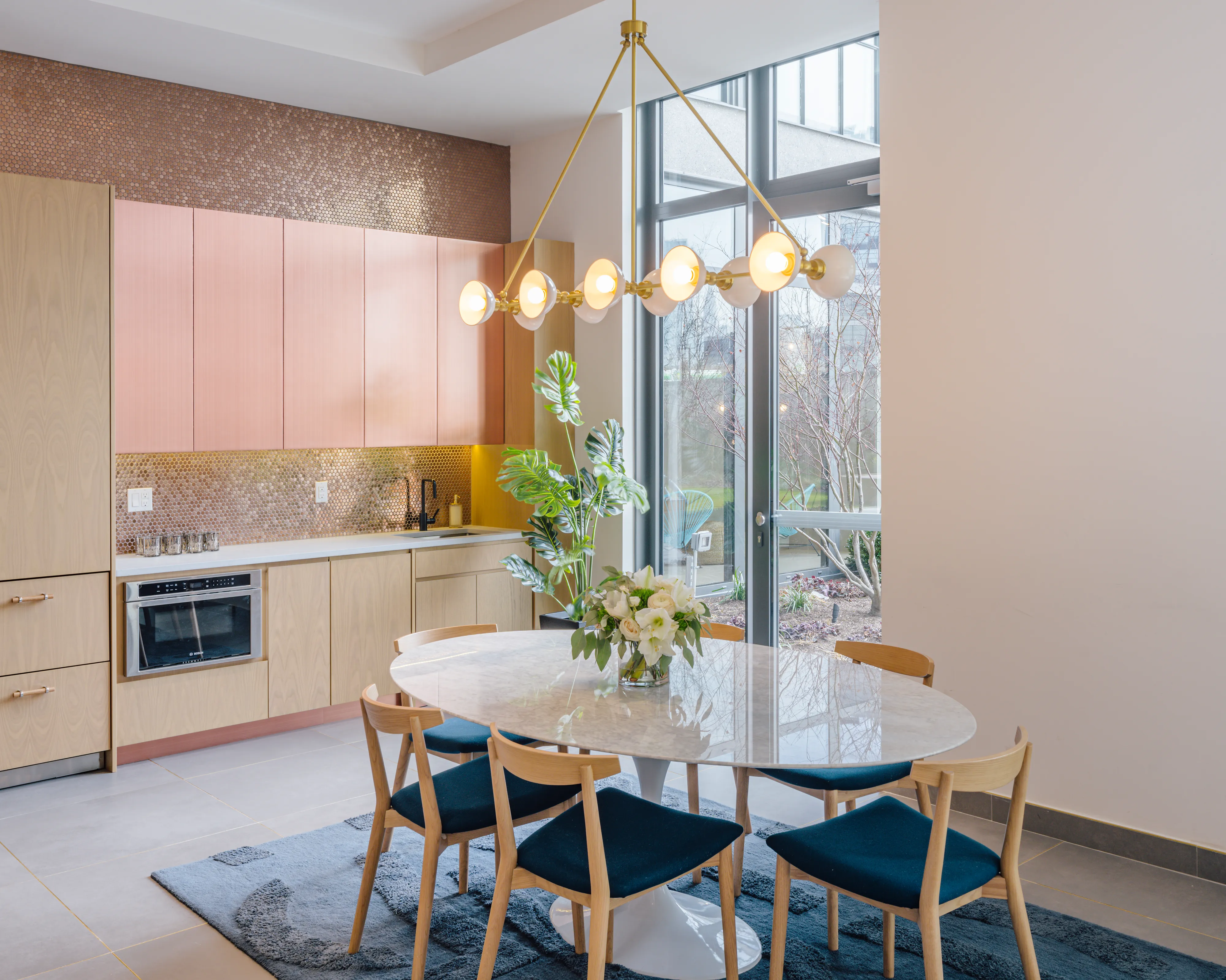 Modern kitchen with pink cabinets, round table, blue chairs, and decorative lighting.