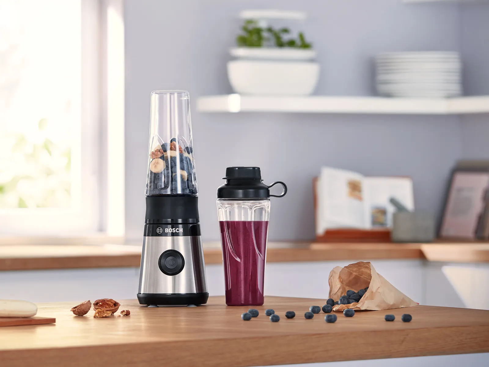 Un blender compact cu recipient transparent, plin cu nuci și fructe de pădure, așezat lângă o sticlă cu un smoothie violet, pe blatul de bucătărie