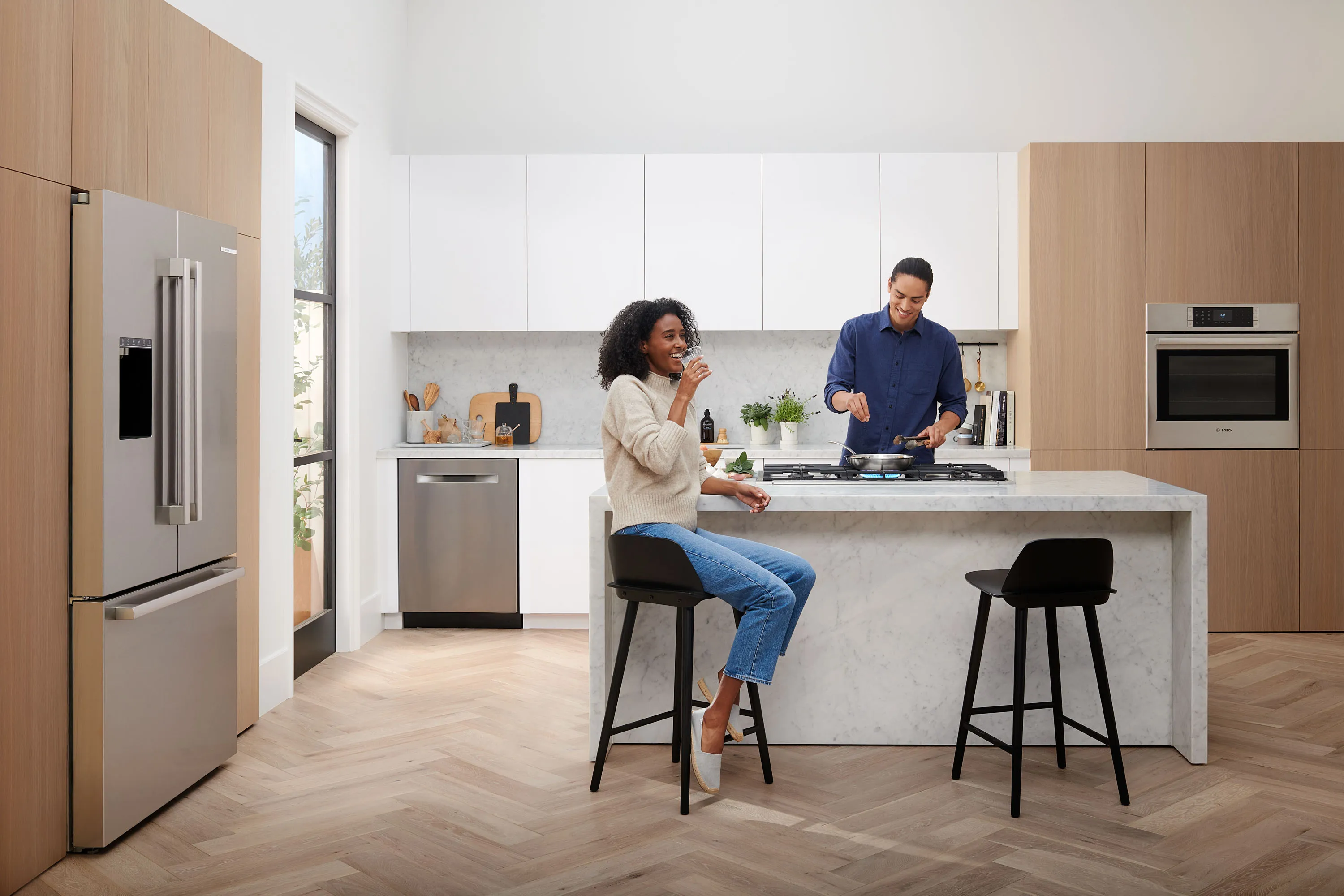 A man cooks on a gas hob on a kitchen island while a woman sits on a bar stool, laughing, in a bright, modern kitchen with Bosch appliances.
