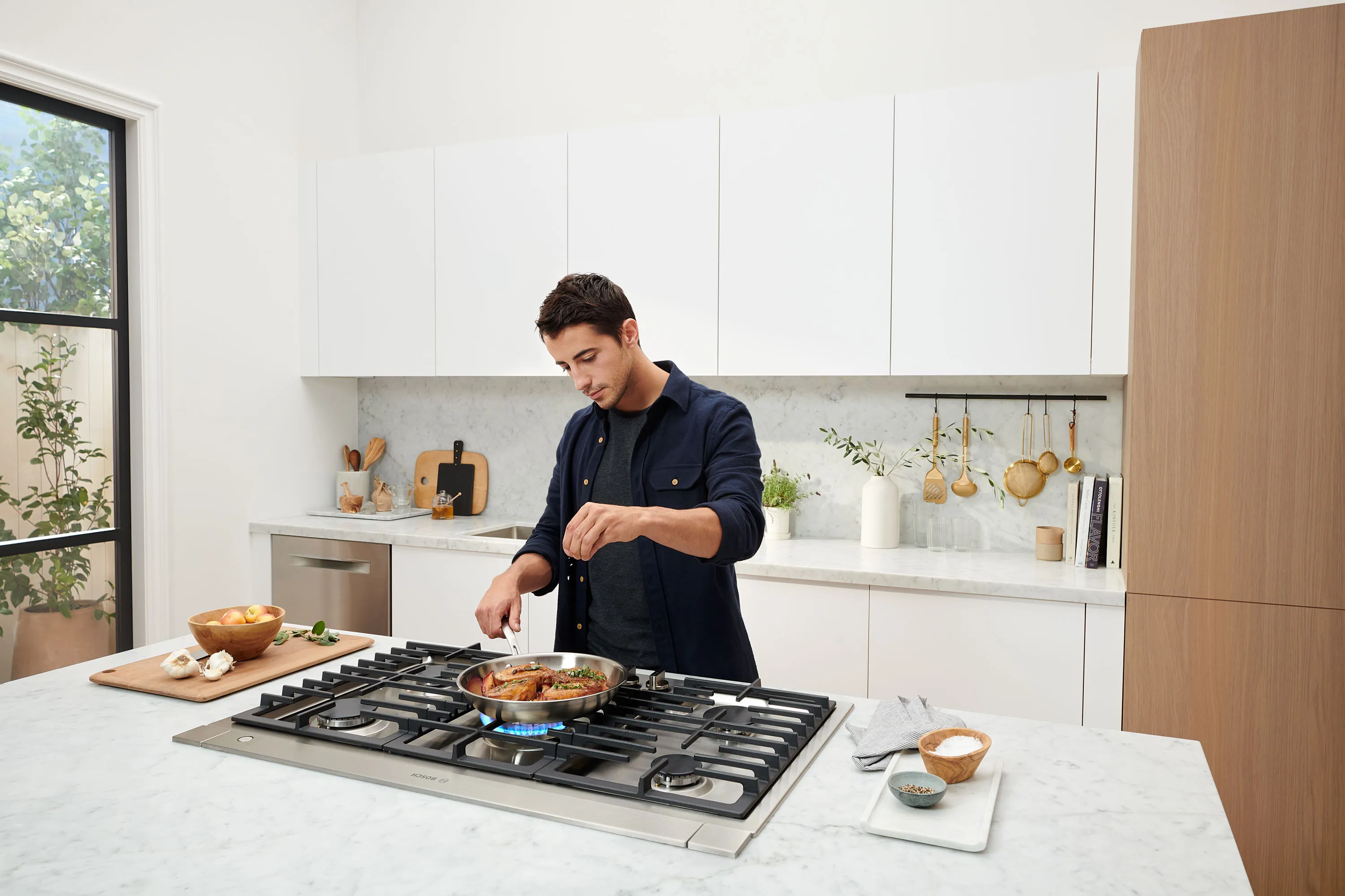 Man in navy blue button up cooking on Bosch gas stove top and seasoning meat cooking in a stainless steel pan