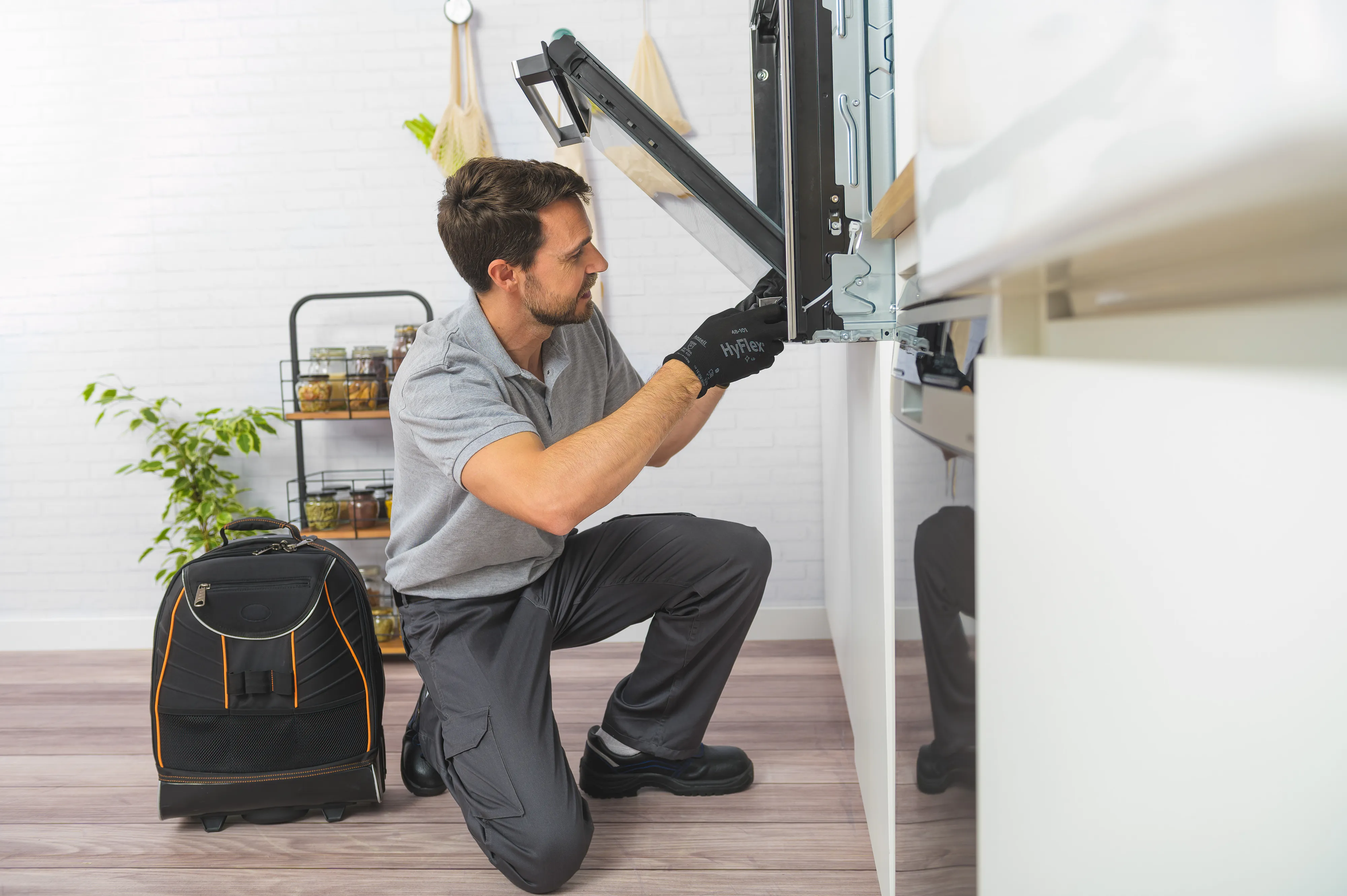 A technician kneels to repair a dishwasher in a bright kitchen, tools nearby.