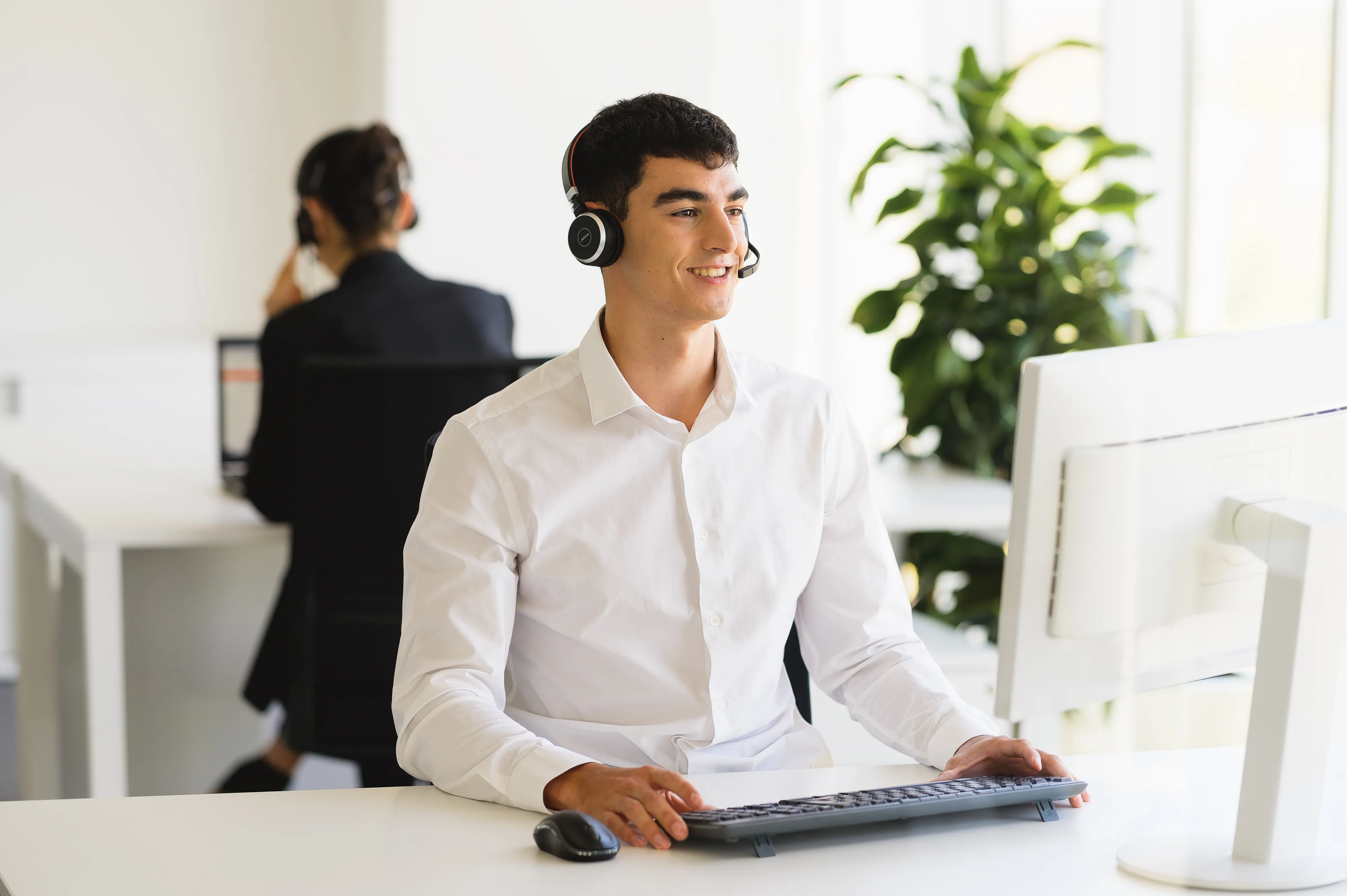 Man wearing headset working at a computer in a modern office with a colleague in the background.