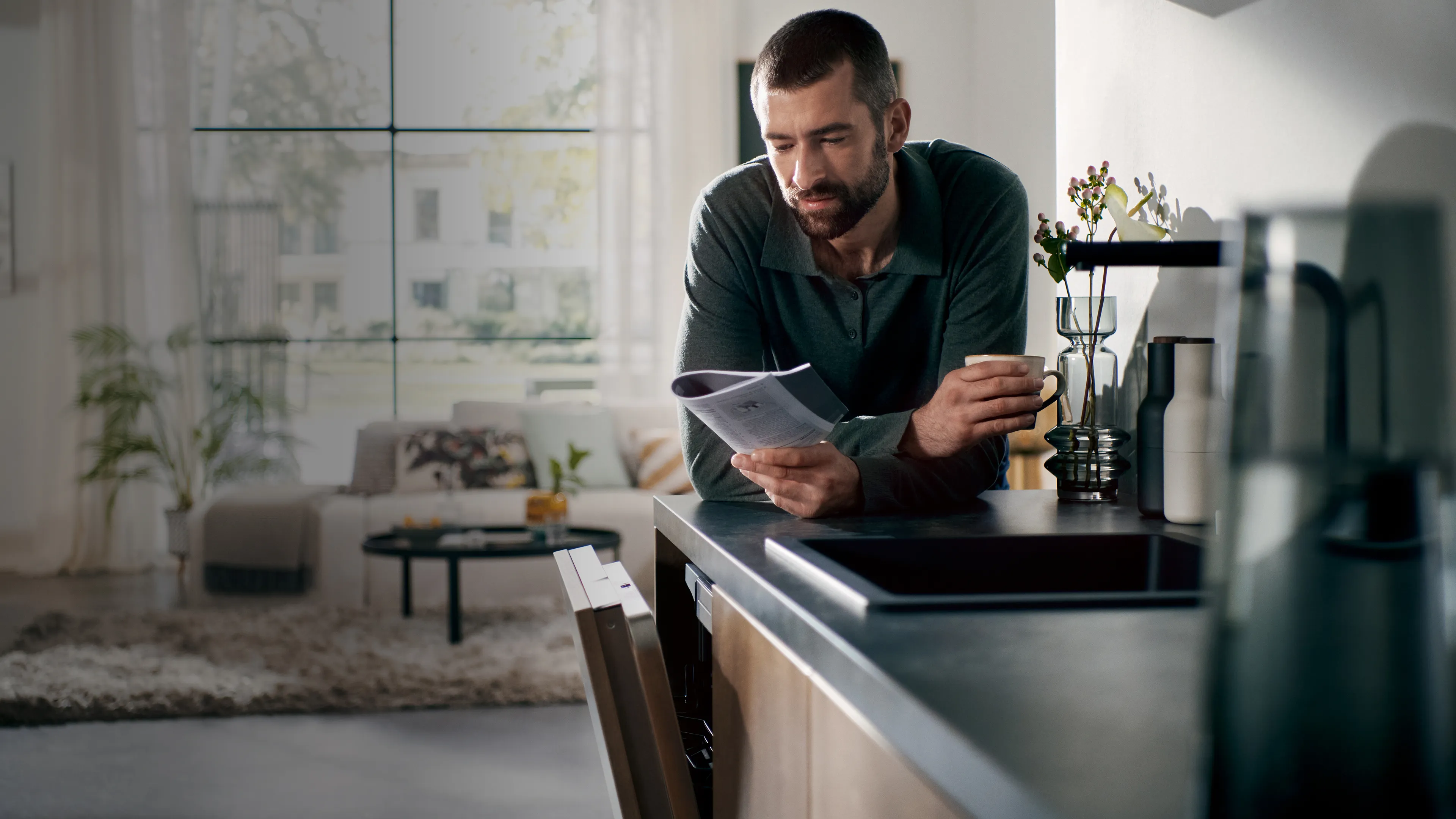 alt-Man reading a user manual while holding a cup in his right hand in a contemporary kitchen, with a view of a bright living room in the background.