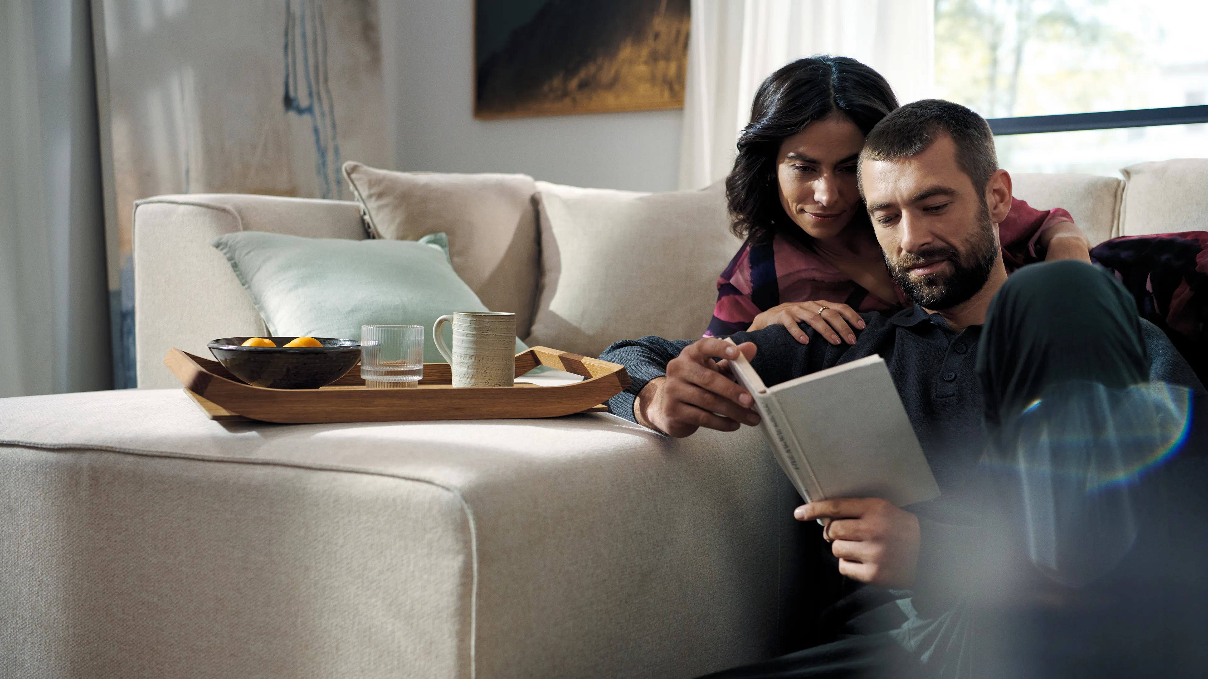 alt-Couple sitting on a beige sofa, with a breakfast tray on the sofa, enjoying reading a book together.