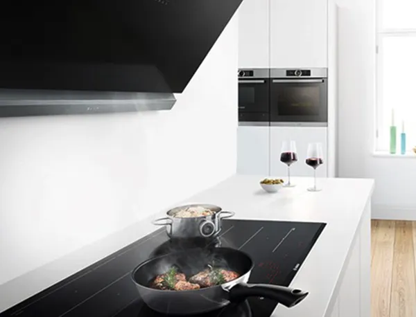 Angled black extractor hood above food cooking on an induction cooktop in a modern white kitchen.