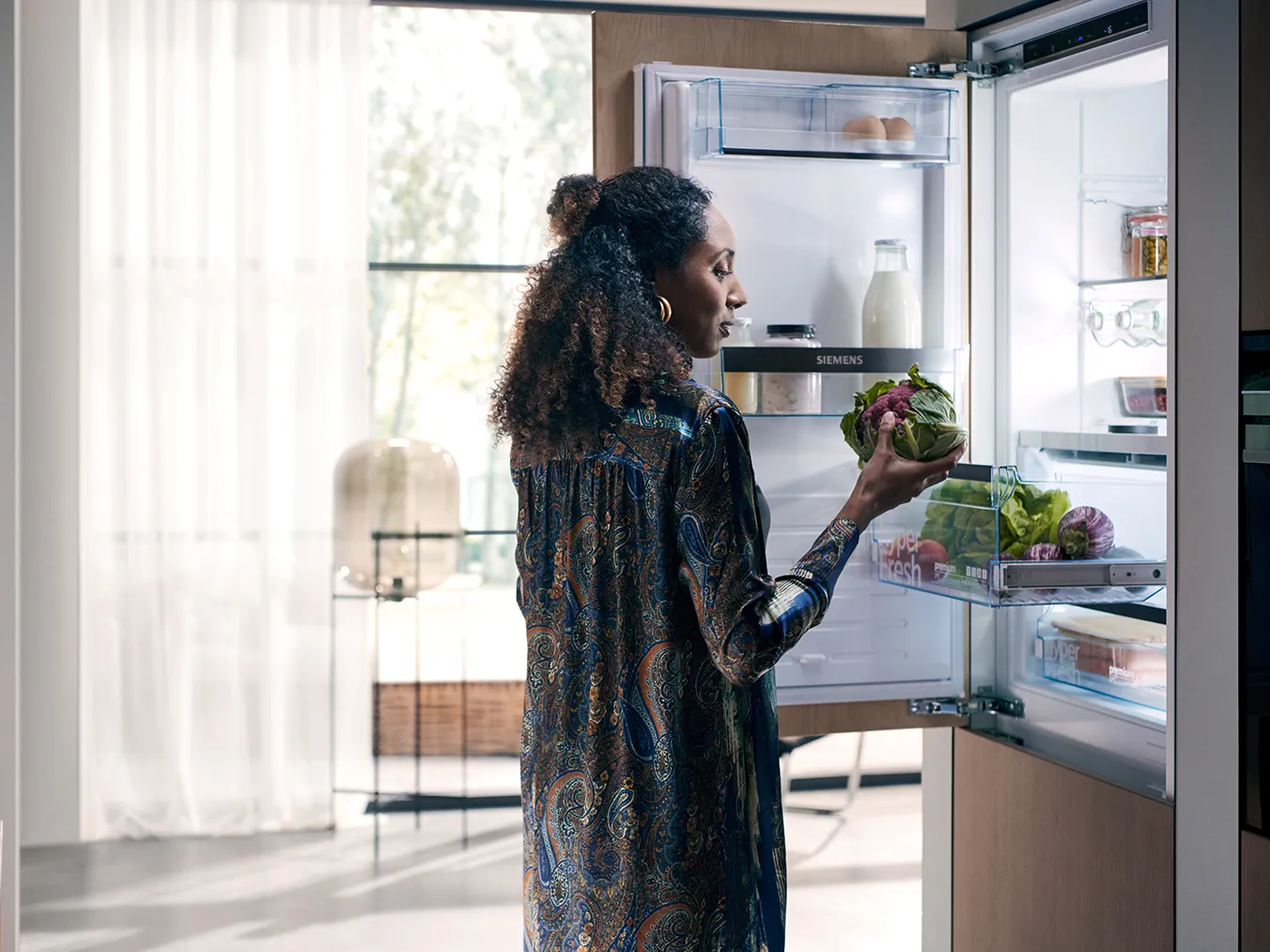 A woman in a patterned robe stands by a modern refrigerator, holding a bowl of fresh vegetables, with bright light streaming in.
