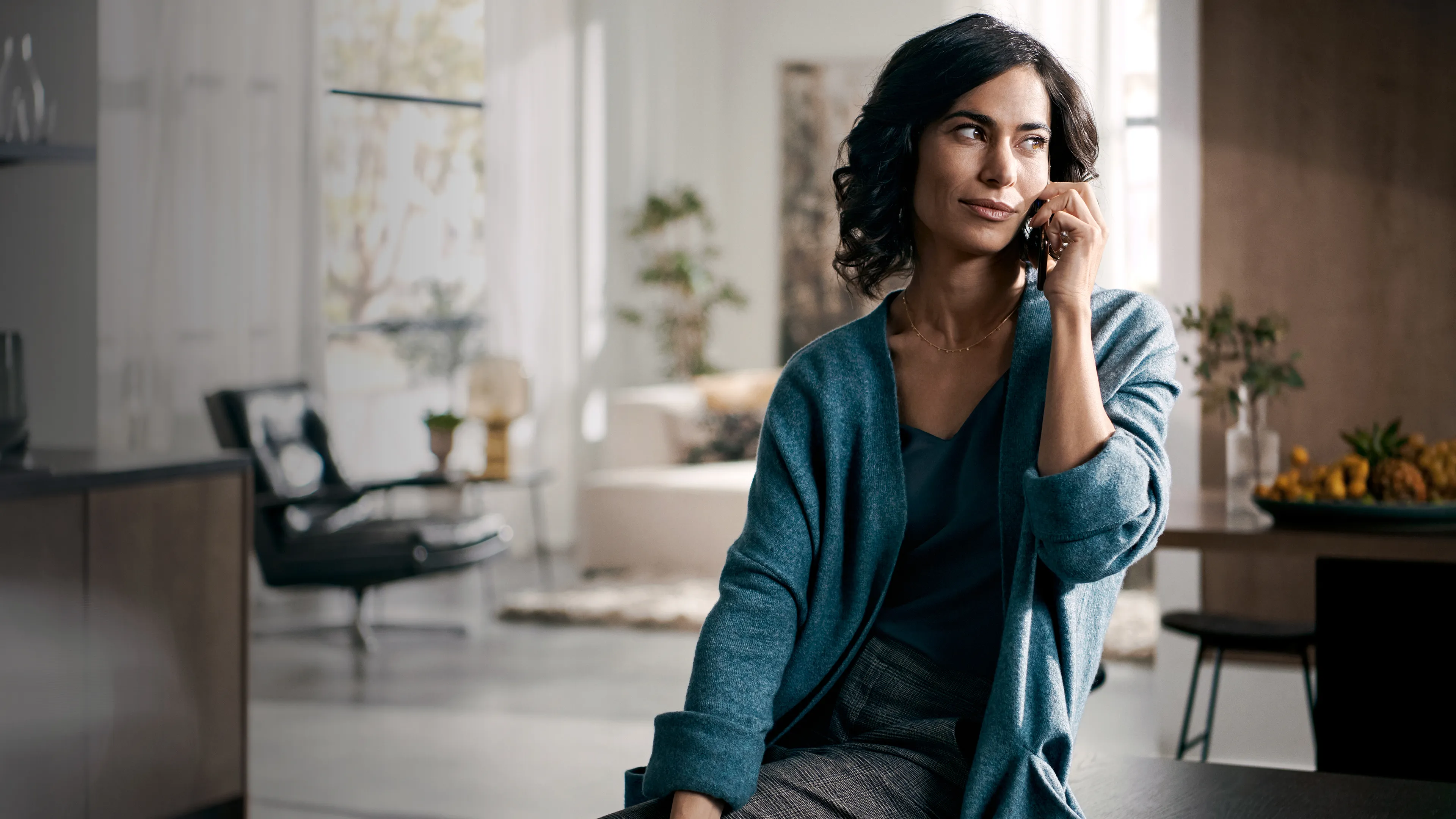 alt-Dark-haired woman with curly hair, wearing a blue cardigan, talking on the phone in a bright, modern space with minimalist furniture.
