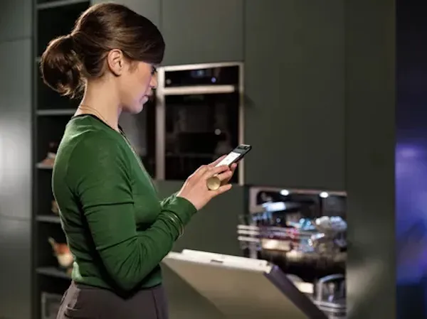 Donna in camicia verde utilizzando smartphone in cucina con elettrodomestici.