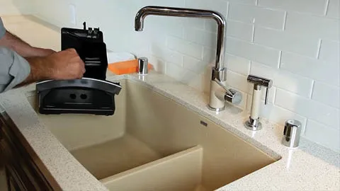 Sink with faucet, hands operating a kitchen appliance, white tile backsplash.