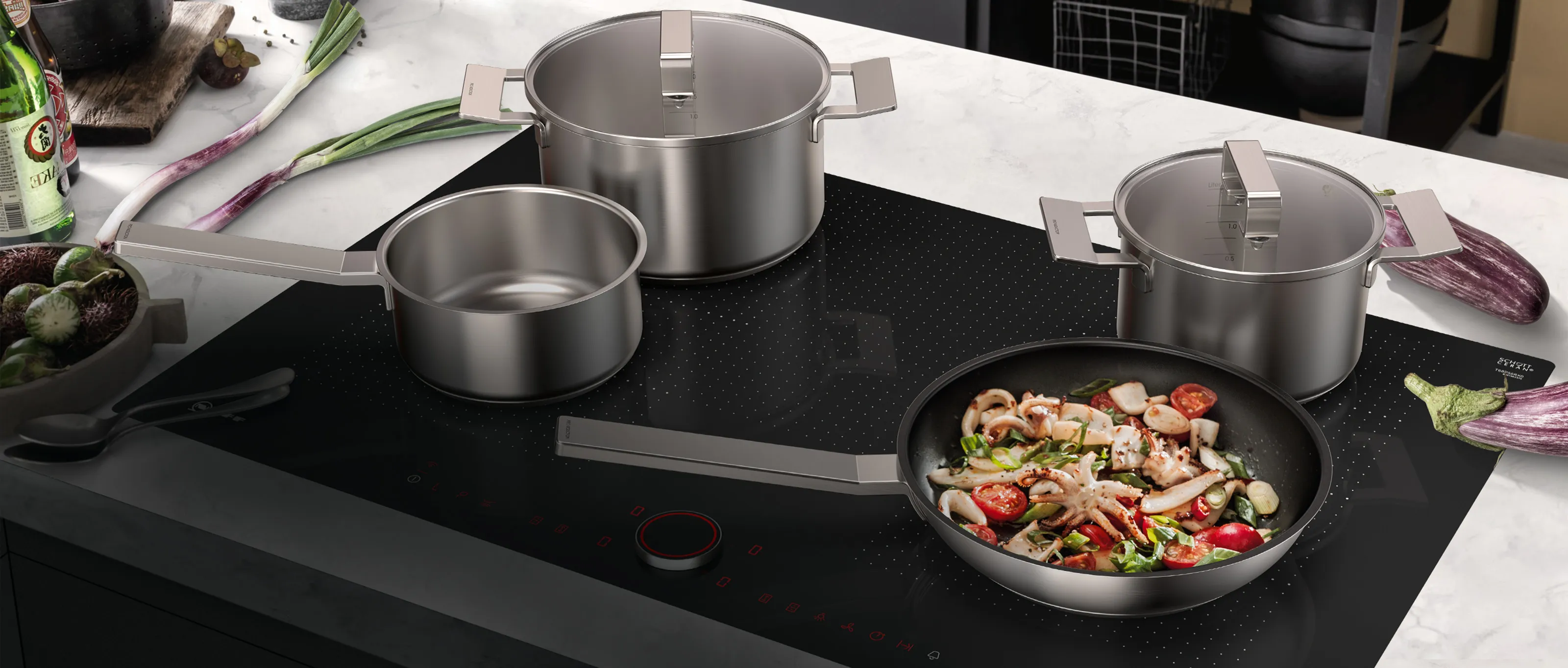 Piano cottura a induzione nero con pentole in acciaio inossidabile e una padella in cui si stanno cucinando frutti di mare e verdure su un piano di lavoro in marmo bianco.