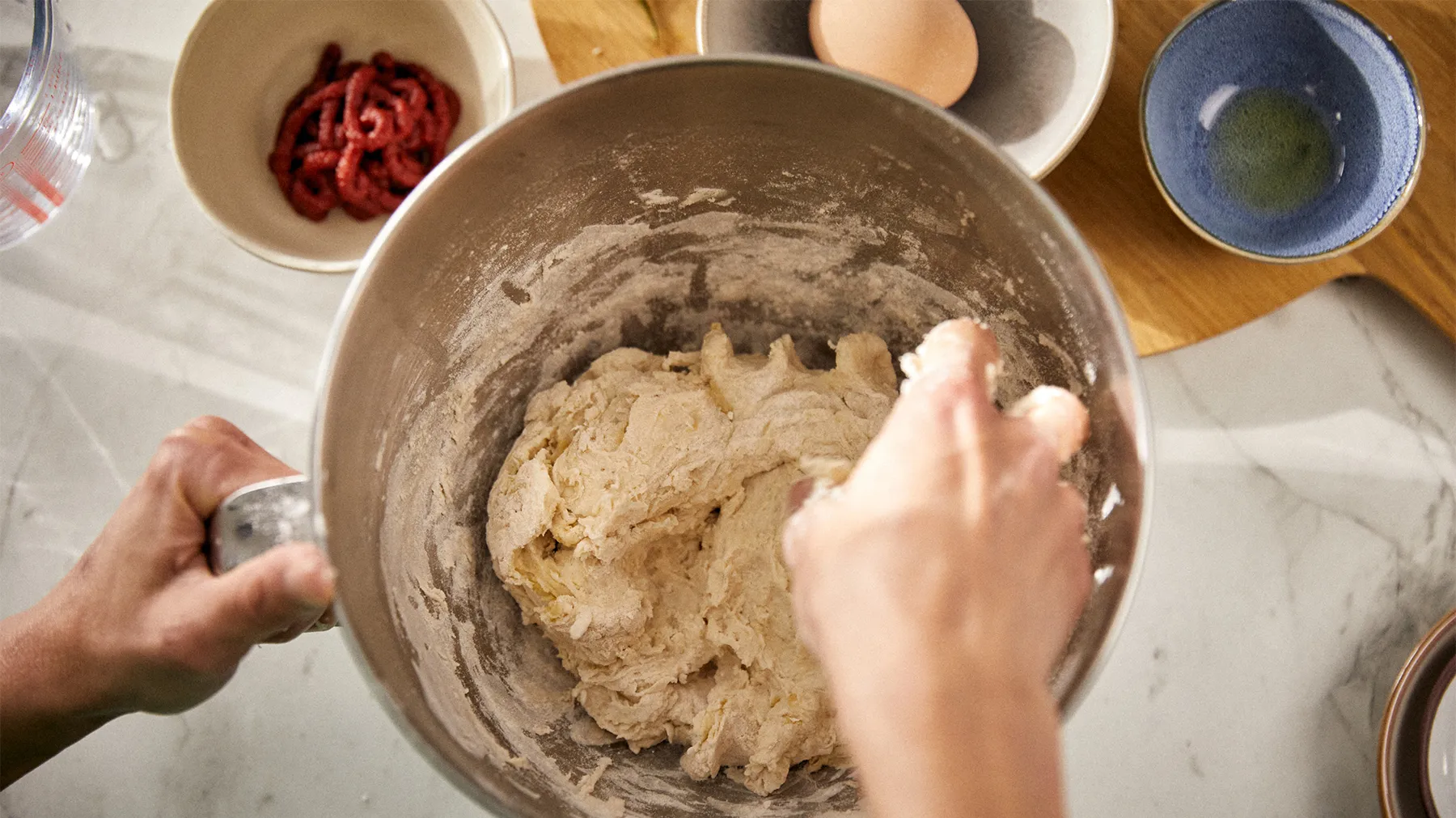 Hands mixing dough in a bowl, surrounded by baking ingredients like eggs and dried fruit.