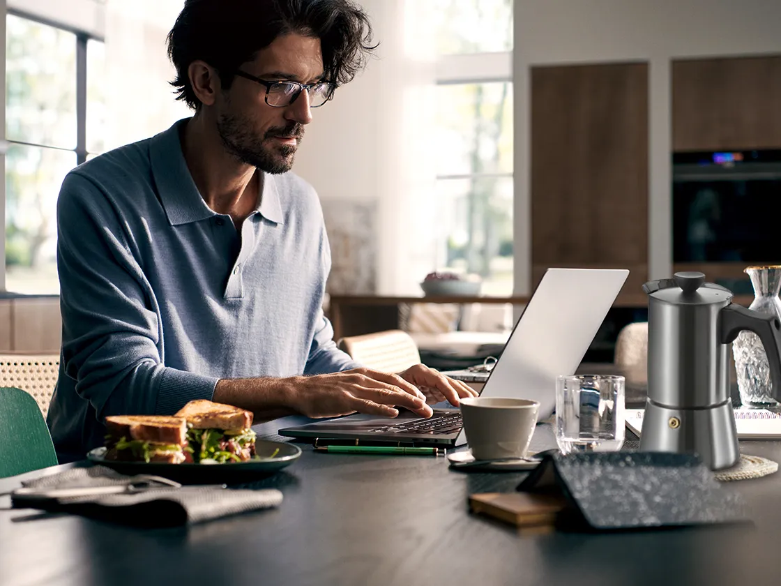 Homem trabalhando no laptop em casa com sanduíche e café na mesa.