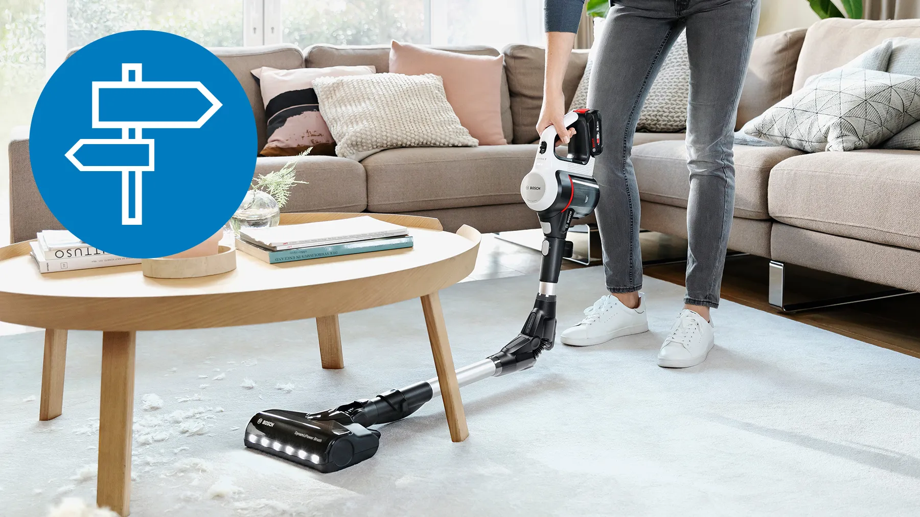 A person using a cordless vacuum cleaner on a white rug, with a directional sign icon in the corner.