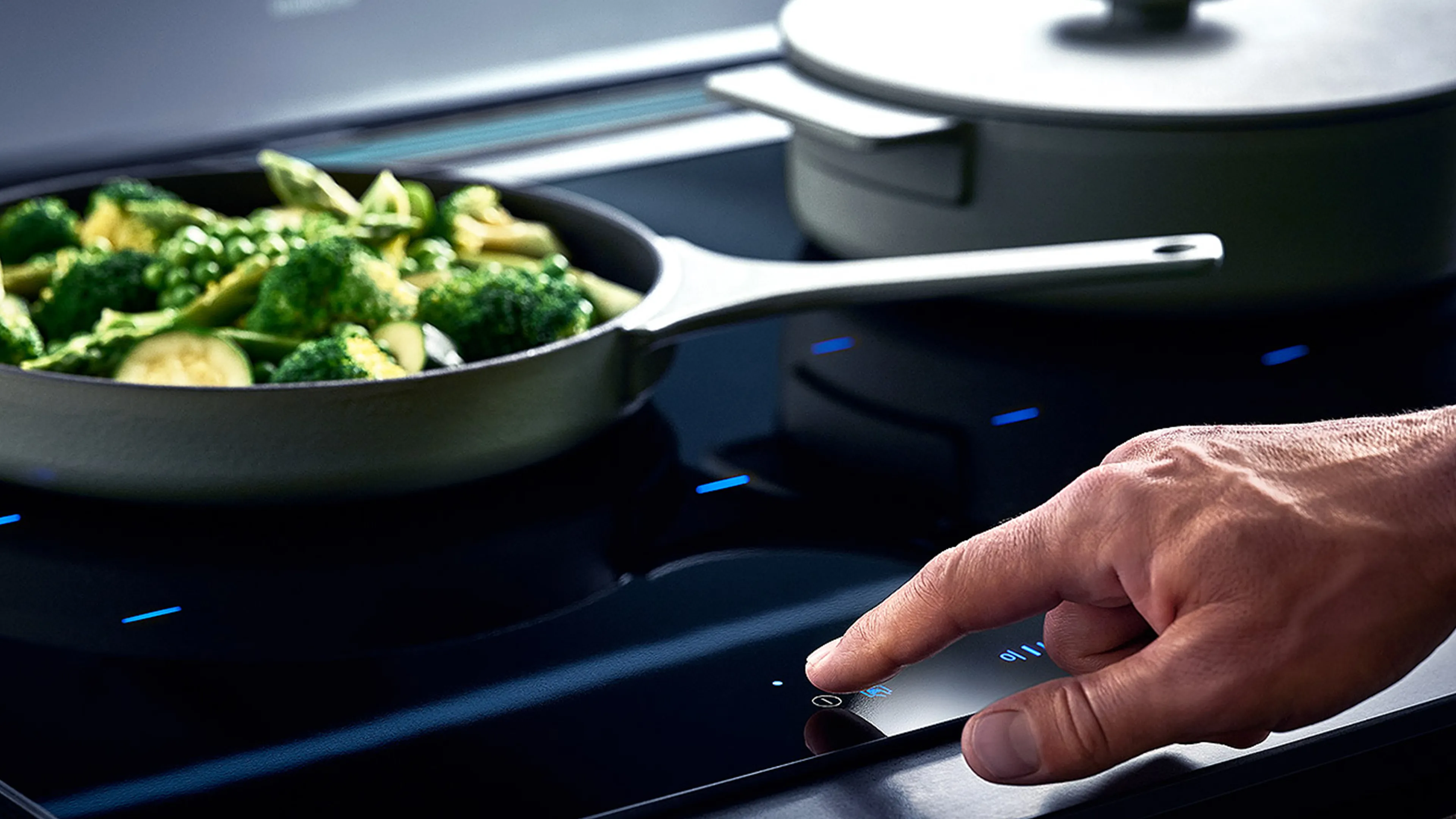 Person using control panel on induction hob whilst a pan of green vegetables is being cooked