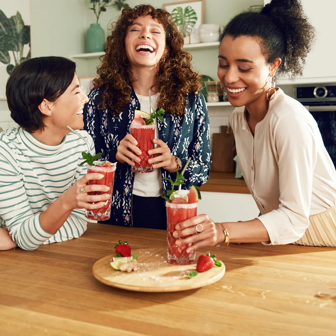Three women holding glasses of red smoothies garnished with mint, strawberry, and lemon, standing around a wooden table with ingredients.