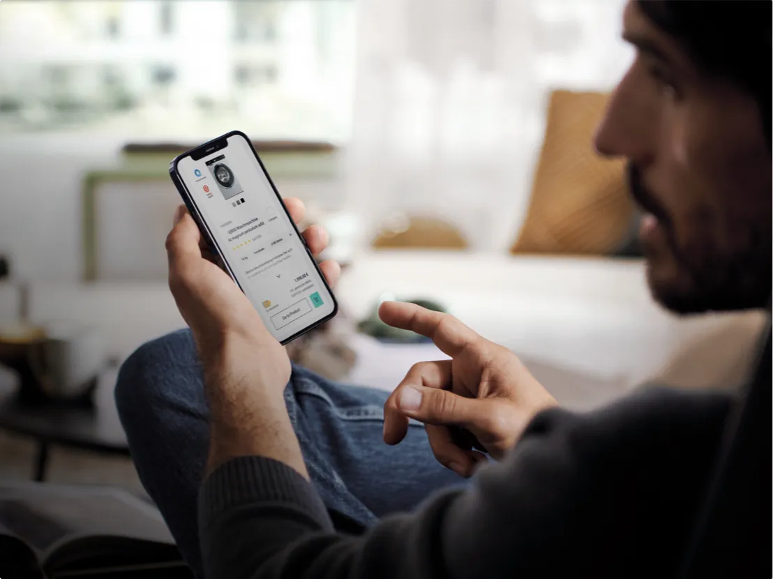 En person håller en smartphone som visar en app, sittande bekvämt i en soffa i ett mysigt vardagsrum.