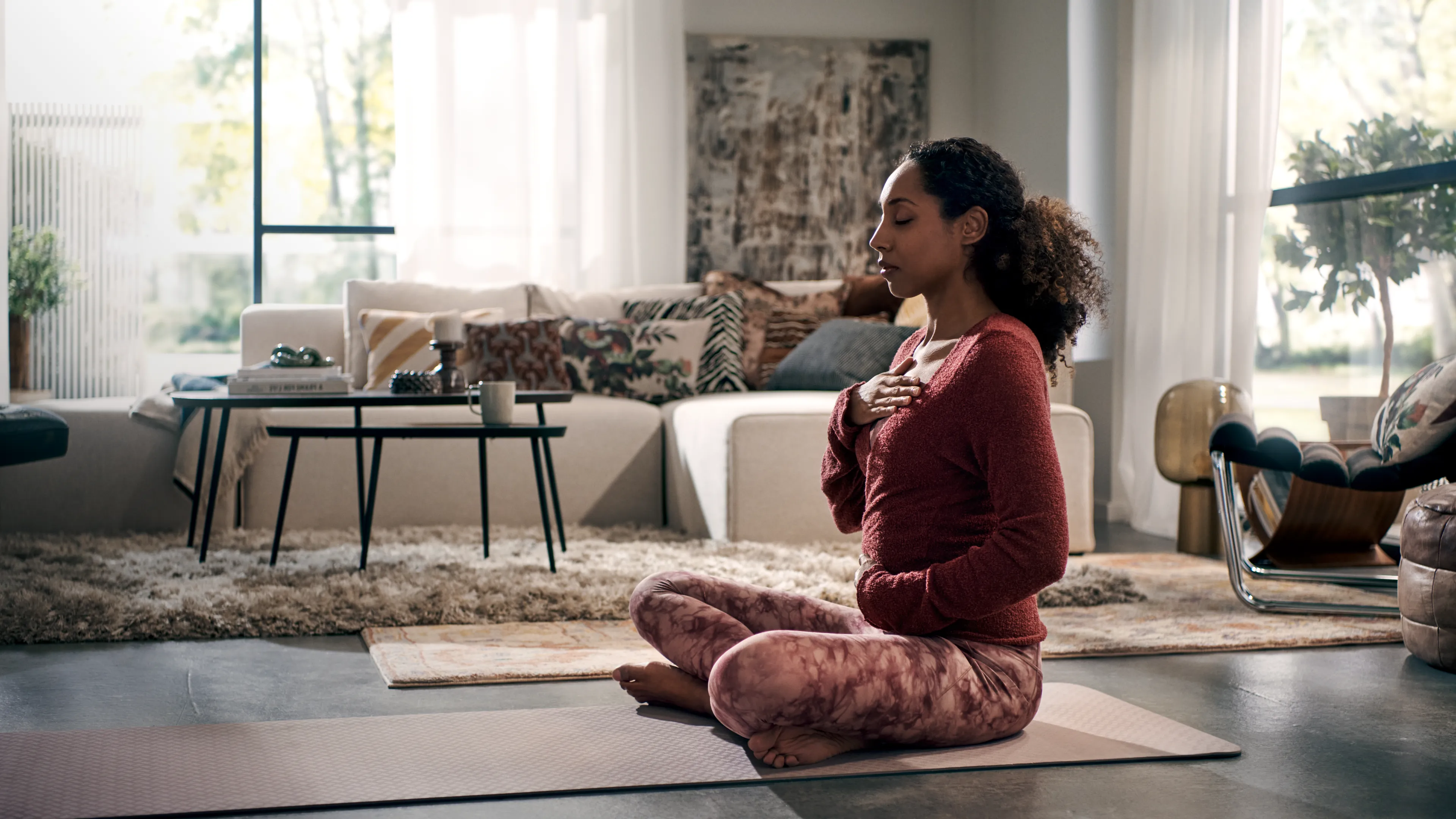 A woman sits cross-legged on a yoga mat in a bright, modern living room, with her eyes closed in a meditative pose. She is wearing a red sweater and patterned leggings, and her hands are placed gently on her chest and stomach. The room is decorated with a comfortable white sofa, plush rugs, and large windows that let in natural light.