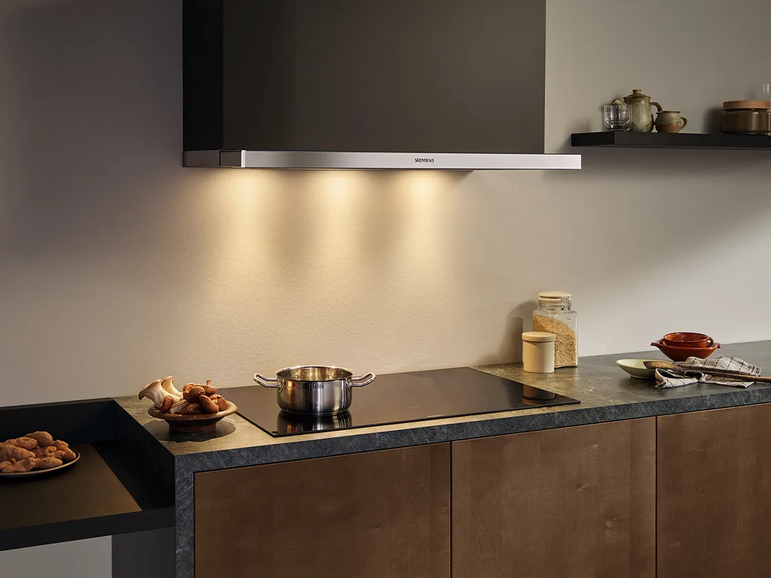 A modern kitchen with a Siemens wall-mounted cooker hood above an induction hob with a pot and a pan of food, and a counter with various kitchen items and ingredients
