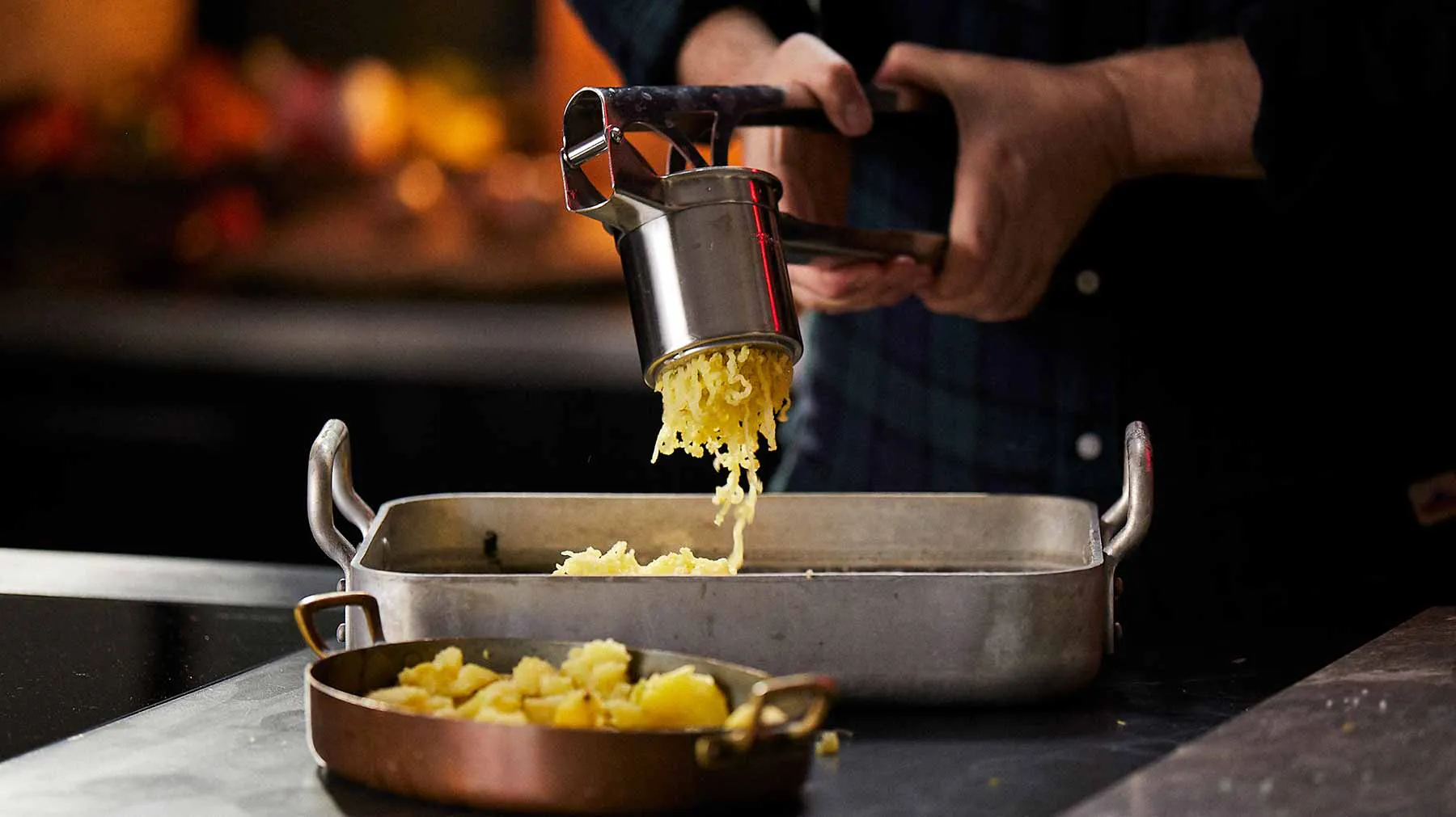 Hands grating cheese into a baking dish with cooked potatoes, creating a cheesy potato dish.