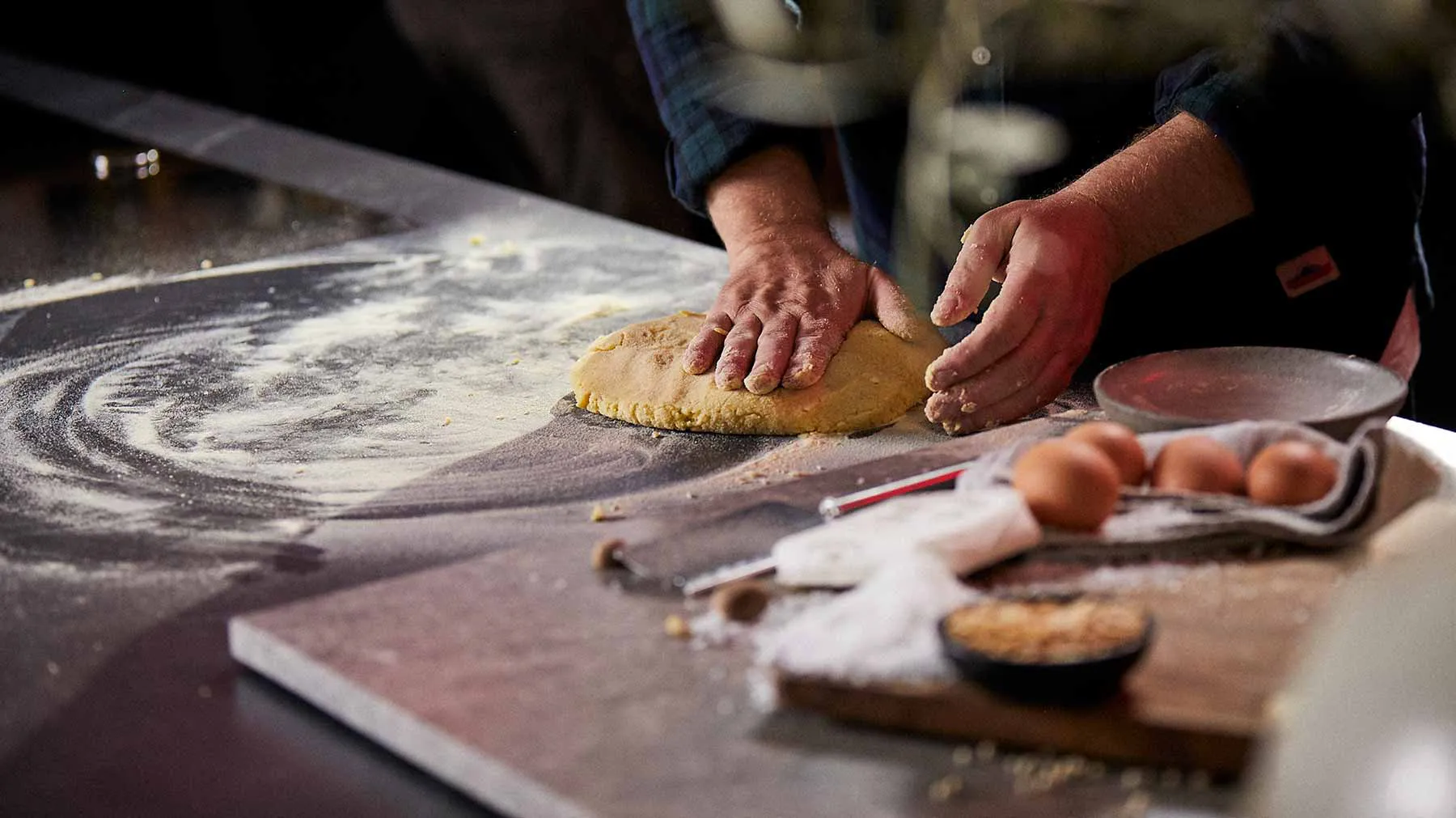 Hands kneading dough on a floured surface, with various baking tools and ingredients visible.