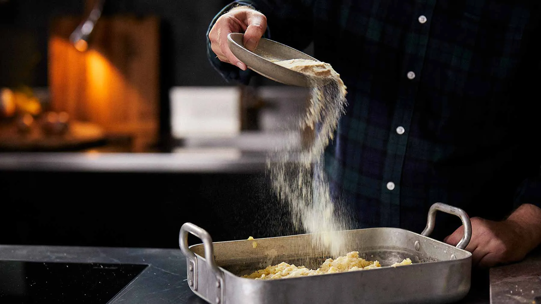 Chef sprinkling seasoning over a dish in a cooking pan on a kitchen counter.
