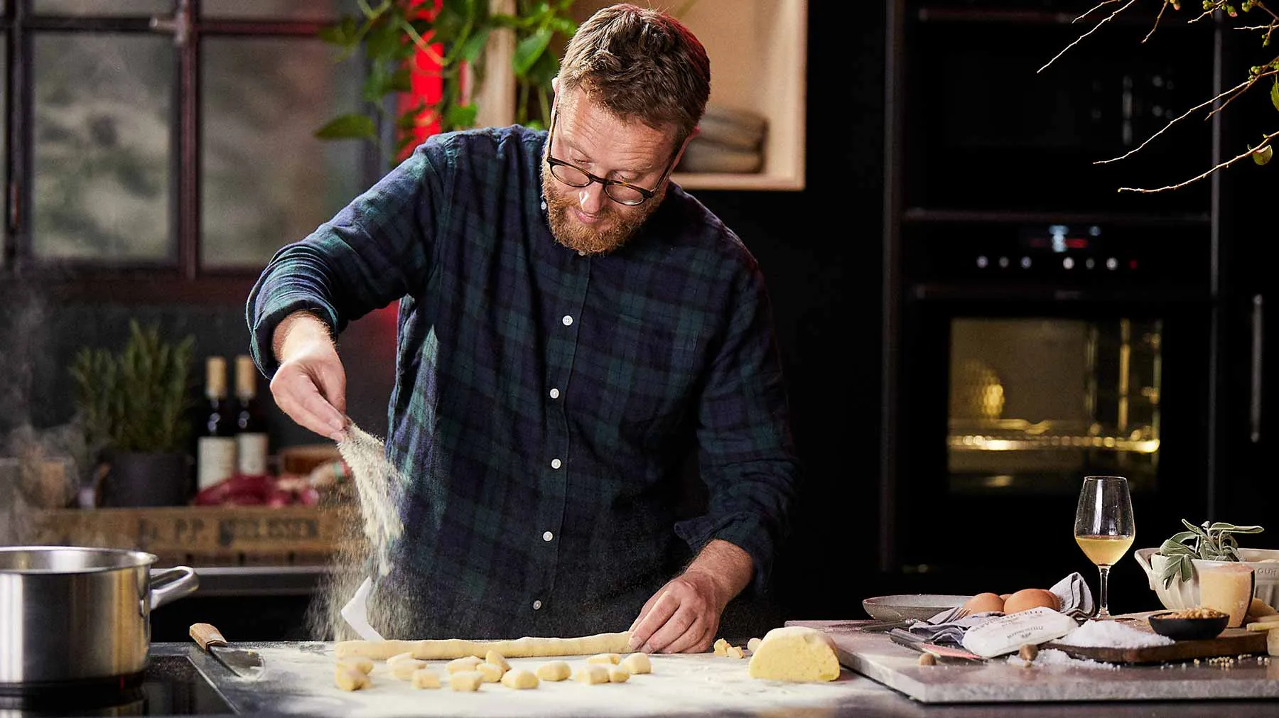 Man with beard cooking in kitchen, preparing food on counter, wine glass on table.